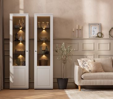 Wohnzimmer in "Cosy Beige" im Landhaus-Stil mit Sofa, beleuchteten Vitrinen und hohen Fenstern.