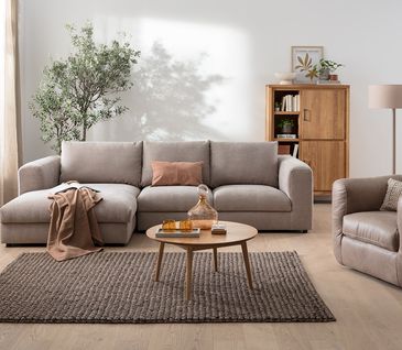 Helles, natürlich eingerichtetes Wohnzimmer mit grauem 3-Sitzer Sofa, braunem Ledersessel und Highboard aus Holz.