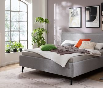 Für das erste eigene Bett kann es schon mehr als 90 Zentimeter Breite sein. Hellgraues Sofa in modernem Schlafzimmer vor fliederfarbener Wand mit Beistelltischen und Highboard.