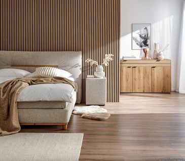 Beiges Boxspringbett in Schlafzimmer vor Holzlamellenwand und Sideboard aus Massivholz.
