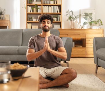 Junger Mann sitzt im Schneidersitz in natürlich eingerichtetem Wohnzimmer und entspannt sich.
