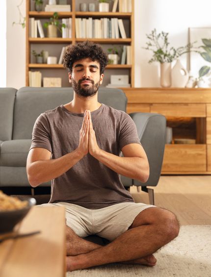 Junger Mann sitzt im Schneidersitz in natürlich eingerichtetem Wohnzimmer und entspannt sich.