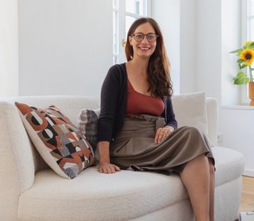 Nicole Zweig sitzt auf einem beigem Bouclé-Sofa mit organischen Formen, dekoriert mit Kissen.