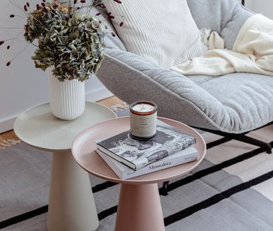 Hellgrauer Sessel mit Kissen und Decke neben zwei runden Beistelltischen.