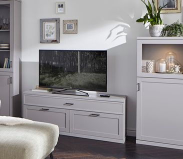 Hellgraue Anrichte neben Lowboard und Fernseher im Landhaus-Look mit beleuchteten Einlegeböden