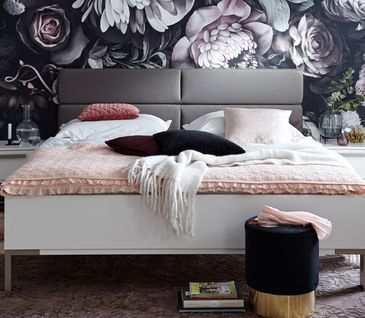 Weißes Bettgestell mit grauem Kopfteil vor bunter Blumentapete