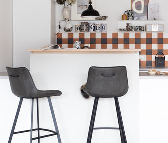 Frontale Sicht auf Küchentresen mit zwei grauen Barhockern und Küchenzeile im Hintergrund mit Wandfliesen in Ocker, Schwarz und Weiß.