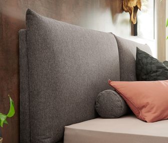 Gepolsterte Rückenlehne eines Betts mit anthrazit-farbenem Stoffüberzug und peach-farbenem Kissen