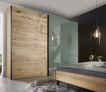 Schrank mit Schwebetür aus Holz und grüngrauen Glas vor einem Massivholzbett