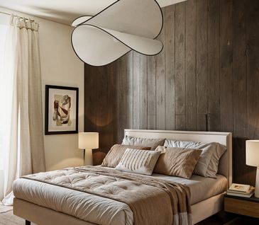 Schlafzimmer mit beigem Boxspringbett vor dunkler Holzwand und auf dunkelbraunem Teppich.