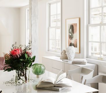 Helles Esszimmer mit weißem Tisch, passenden Stühlen und Sideboard. Dekoriert mit Blumen und Vasen.