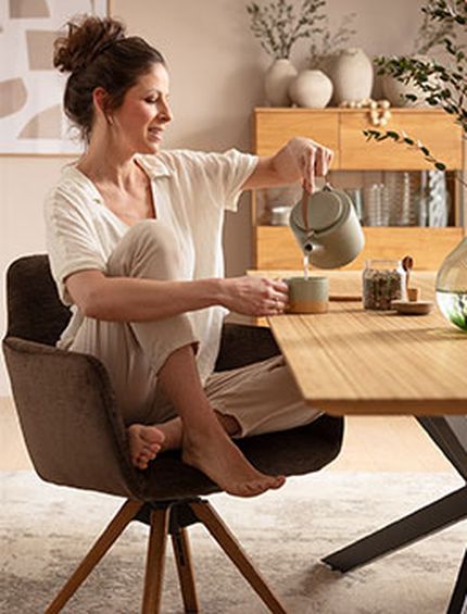 Frau genießt ihren Tee beim Frühstück am Esstisch auf bequemen Armlehnstuhl mit Holzgestell.