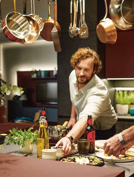 Dein Zuhause liebt dich - Sichern Sie sich jetzt Ihre neue Contur Küche zu exklusiven Vorteilen bei teilnehmenden Händlern. Zwei Männer kochen in moderner Küche mit roten Fronten. Töpfe hängen von oben herab.