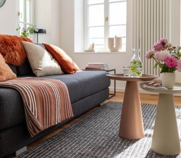 Graues Sofas, dekoriert mit Decken und Kissen neben zwei Beistelltischen in Beige und Rosa.