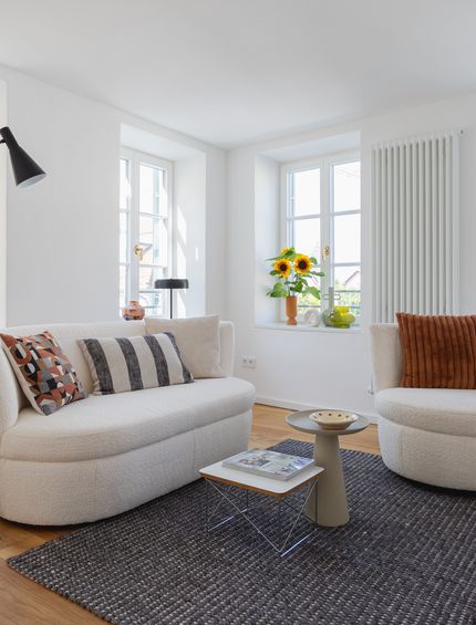 Helles Wohnzimmer in Altbauwohnung mit beigem Sofa und Sessel in organischen Formen, Beistelltischen, schwarzer Lampe und Kissen dekoriert.