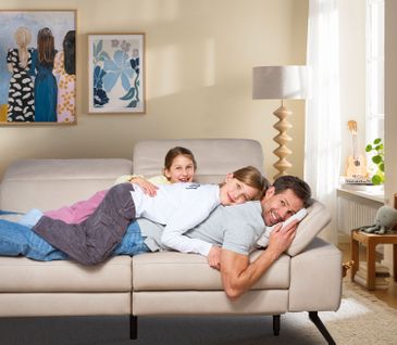 Vater liegt entspannt mit zwei jungen Töchtern auf gemütlichem Sofa in hellem Wohnzimmer.