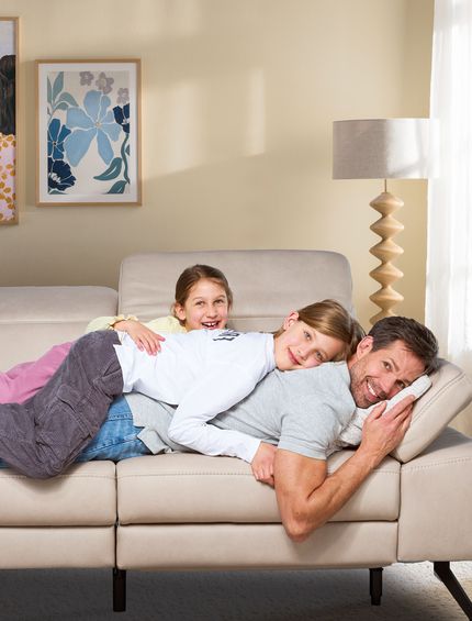 Vater liegt entspannt mit zwei jungen Töchtern auf gemütlichem Sofa in hellem Wohnzimmer.