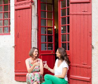 Zwei Frauen sitzen entspannt auf den Stufen vor einem Hauseingang mit roten Fensterläden.