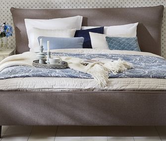 Graues Polsterbett dekoriert mit vielen Kissen und einer Tagesdecke in weiß und Blautönen. Beistelltisch mit moderner Lampe.