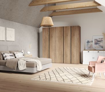 Gemütliches Schlafzimmer mit hellgrauem Boxspringbett, rosa Sessel, weißem Sideboard und großem Holzschrank.