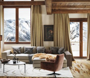 Wohnzimmer im Chalet-Stil mit grauem Ecksofa, braunem Drehsessel, vielen Kissen und Decken mit Aussicht auf die Berge.