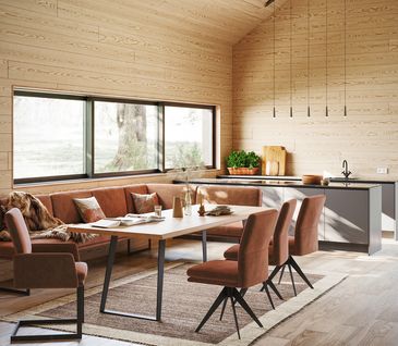Offene Wohnküche mit Küchenzeile und Essbereich im Chalet-Stil mit holzverkleideten Wänden, Holz-Esstisch und Eckbank mit Stühlen in der Farbe Cognac.