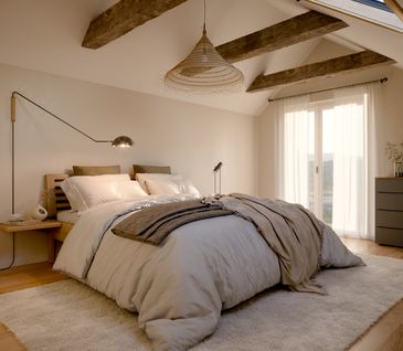 Hell eingerichtetes Schlafzimmer mit beiger Tagesdecke, Teppich und grauer Kommode.