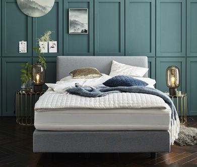 Frontansicht auf Boxspringbett mit Decke und verschiedenen Kissen vor türkiser Wand