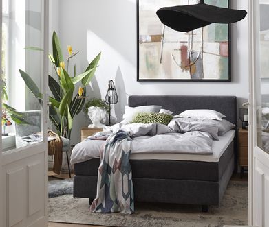 Schlafzimmer mit weißer Wand und geöffneter Doppeltüre. Dunkelgraues Boxspringbett mit hellen Kissen und heller und bunter Decke dekoriert.