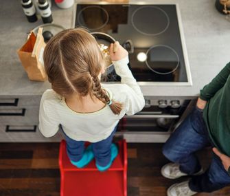 Mädchen steht mit Elternteil am Herd und kocht