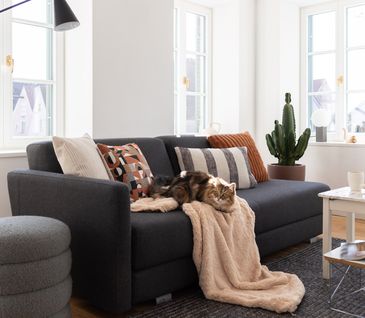 Helles Wohnzimmer mit grauem Sofa, dekoriert mit vielen Kissen und einer beigen Decke.