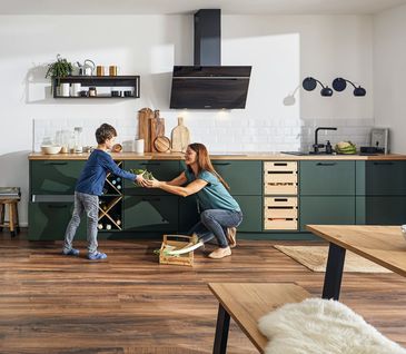 Frau reicht ihrem Sohn Gemüse vor einer grünen Küche