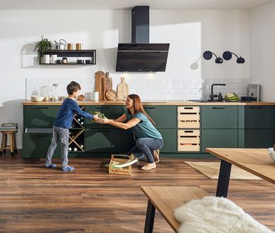 Frau reicht ihrem Sohn Gemüse vor einer grünen Küche