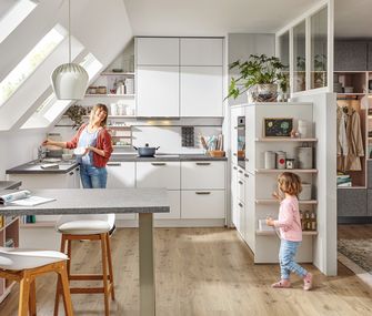 Frau kocht in weßer Küche im Dachgeschoss und bekommt Gesellschaft ihrer kleinen Tochter