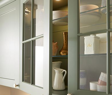 In grünen Küchenschränken mit Glastüren steht weißes Geschirr. Sanfte Beleuchtung unterstreicht das elegante Arrangement.