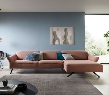 Rosanes Stoffsofa vor hellblauer Wand mit grauem Teppich und kleinem Beistelltisch. Dekoriert mit bunten Kissen.