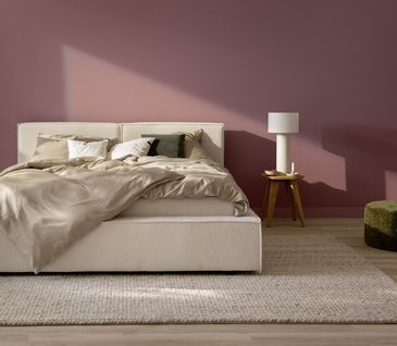 Schlafzimmer mit Wand in Rosé und beigem Bett mit Beistelltisch und grünem Hocker.