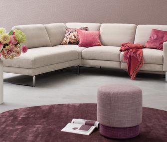 Beiges Ecksofa mit Kissen in Rosa und Pink vor lila Teppich mit lila Hocker.