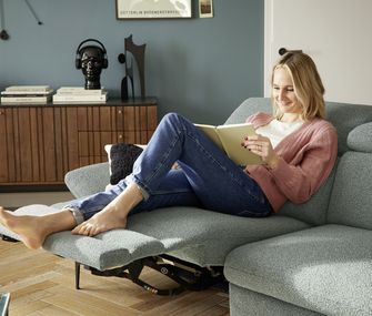 Frau liest auf einem Sofa mit Relaxfunktion ein Buch und legt dabei die Beine hoch.