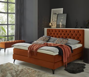 Orangefarbenes Boxspringbett mit Topper, Bettbezug und passender Polsterbank vor Fenster.