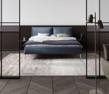 Blaues Bett vor weißer Wand mit holzfarbener Verkleidung. Blaue Tagesdecke auf Bett mit hellen Kissen.