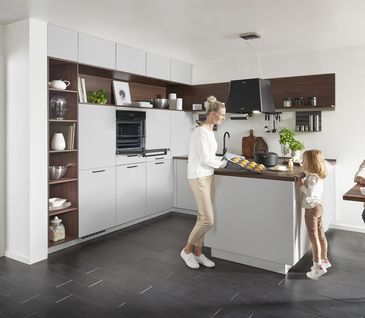 Familie in einer grauen Eckkueche mit Holzakzenten beim gemütlichen Frühstück im Essbereich und Backen im hochgelagerten Backofen.