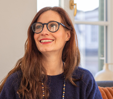 Nicole Zweig lächelt und sitzt auf gemütlichem Sofa.