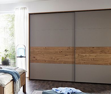 Schrank mit Schwebetür aus Holz und grüngrauen Glas vor einem Massivholzbett