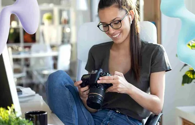 Smiling woman in glasses and earbuds reviews photos on a DSLR camera in a bright office, with decorative purple and blue shapes.