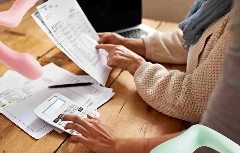 Two people review bills, one pointing at a document while another uses a calculator on a wooden table with a laptop.