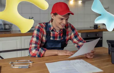A smiling male worker in a red cap and overalls reviews papers at a kitchen table with tools. Abstract yellow and blue shapes float.