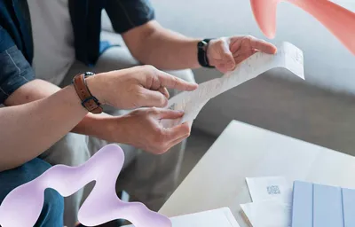 Two people examine a long paper receipt, one pointing at it, with other documents and a tablet visible on a table.