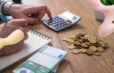 Hands using a calculator and writing in a notebook, with Euro banknotes and coins on a wooden desk, signifying financial tasks.