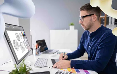 A man in glasses uses a graphics tablet to design intricate coin-like graphics on a computer monitor in a modern office.